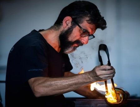Persona trabajando con vidrio caliente, sosteniendo una pieza incandescente con unas pinzas mientras aplica fuego para moldearla. Se observan llamas y humo saliendo del material, en un entorno interior con iluminación tenue.