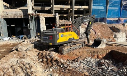 Excavadora de orugas Volvo, color amarillo y negro, con el logotipo de la empresa Benjumea en el costado, trabajando en una obra de demolición y excavación. El terreno está lleno de escombros y tierra removida, y al fondo se observan estructuras parcialmente demolidas y edificios en construcción con andamios y lonas azules.