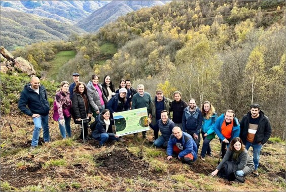 Grupo de personas posando en una ladera con vegetación y montañas al fondo, junto a un cartel informativo sobre reforestación