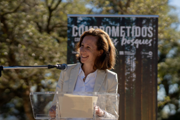 Persona hablando en un atril con micrófono durante un evento al aire libre, con un cartel de fondo que dice 'Comprometidos con los bosques