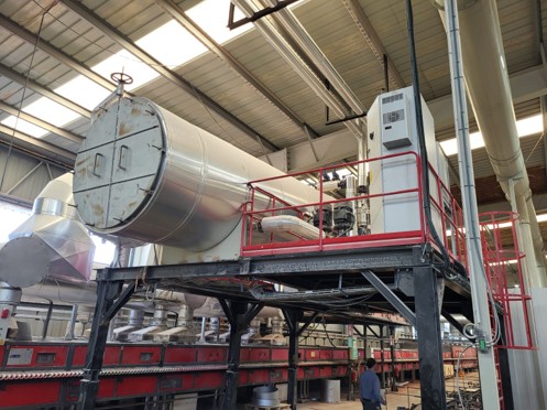 quipo industrial elevado sobre una estructura metálica con barandillas rojas en el interior de una nave. El sistema incluye un gran tanque cilíndrico horizontal de acero inoxidable, conectado a tuberías y componentes de control. A la derecha del tanque hay un cuadro eléctrico vertical. El techo de la nave es metálico con paneles translúcidos que permiten la entrada de luz.