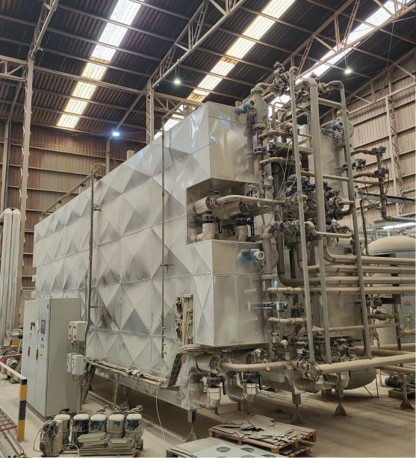 Equipo industrial de gran tamaño instalado en el interior de una nave, con estructura metálica recubierta por paneles de acero inoxidable con diseño geométrico. El sistema incluye múltiples tuberías, válvulas y componentes de control, así como un cuadro eléctrico en la parte frontal. El techo de la nave presenta vigas metálicas y paneles translúcidos que permiten la entrada de luz.