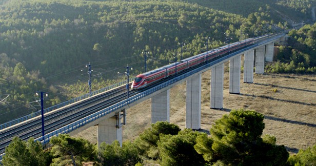 La imagen muestra un tren de alta velocidad circulando sobre un viaducto elevado en una zona montañosa. El tren es de color rojo con detalles en gris y está compuesto por varios vagones aerodinámicos. El viaducto está sostenido por pilares de hormigón altos y atraviesa un paisaje natural con abundante vegetación, principalmente pinos, en el fondo y en las laderas. El cielo no es visible, pero la iluminación indica que es de día, con luz natural clara.