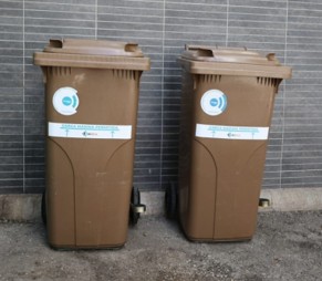 Dos contenedores marrones para residuos orgánicos, colocados uno al lado del otro sobre un suelo de cemento, frente a una pared gris con azulejos. Cada contenedor tiene una etiqueta blanca con un logotipo circular azul y texto que indica “Materia orgánica biodegradable”.