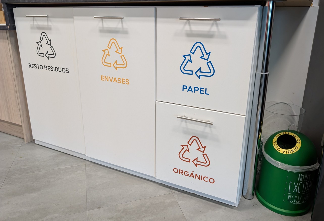 Mueble blanco con cuatro compartimentos para reciclaje: resto de residuos, envases, papel y orgánico, cada uno identificado con símbolo de reciclaje y color distinto. A la derecha, un mini contenedor verde para reciclaje de vidrio