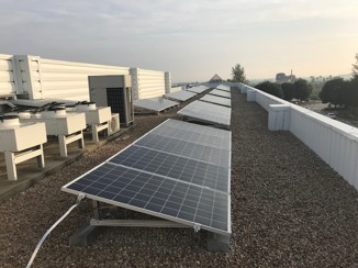 Paneles solares instalados en el tejado de un edificio industrial, colocados en fila sobre una superficie de grava. Al fondo se observan equipos de climatización y un paisaje con árboles y cielo despejado.