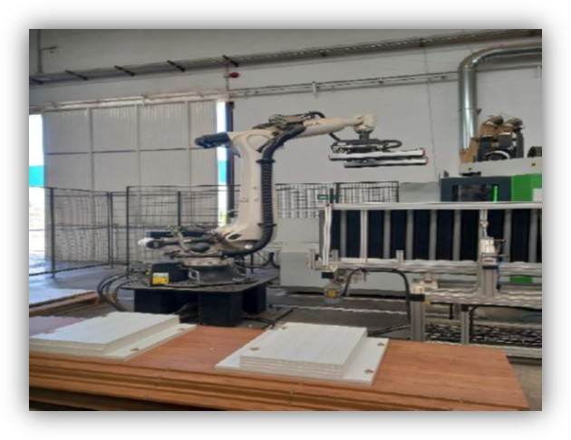 Brazo robótico industrial tomando o colocando piezas rectangulares blancas sobre una mesa de trabajo dentro de una nave de producción, con maquinaria y vallas de seguridad alrededor.