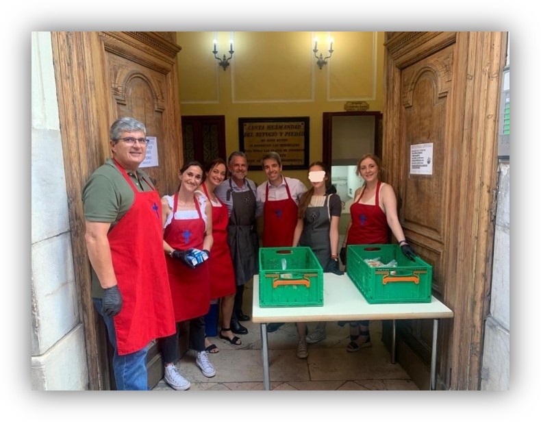 Grupo de personas con delantales junto a una mesa con cajas verdes en la entrada de un edificio