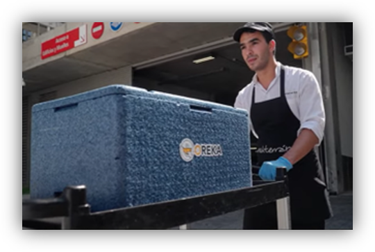 Persona con uniforme manipulando una gran caja isotérmica azul sobre un carrito frente a un acceso de carga