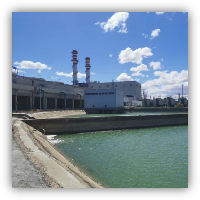 Vista de una planta industrial junto a un canal de agua verde, con dos chimeneas altas al fondo bajo un cielo azul con nubes.