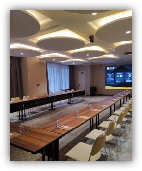 Sala de reuniones preparada para una conferencia, con mesas en forma de U, sillas alineadas y una pantalla grande encendida al fondo.