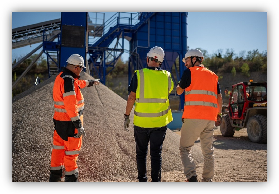 Personas con cascos y chalecos de seguridad supervisan material a granel en una instalación industrial.