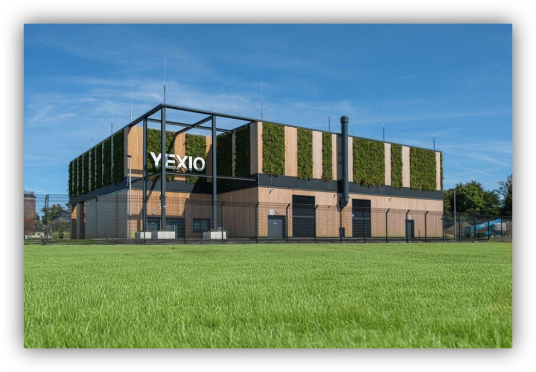 Edificio industrial moderno con fachada vegetal y paneles de madera, rodeado de una zona ajardinada.