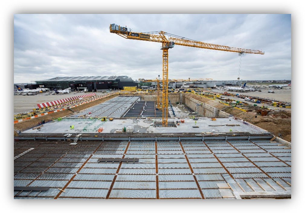 Obras de construcción en un aeropuerto con una gran grúa torre, una excavación de gran tamaño y varias aeronaves visibles al fondo.