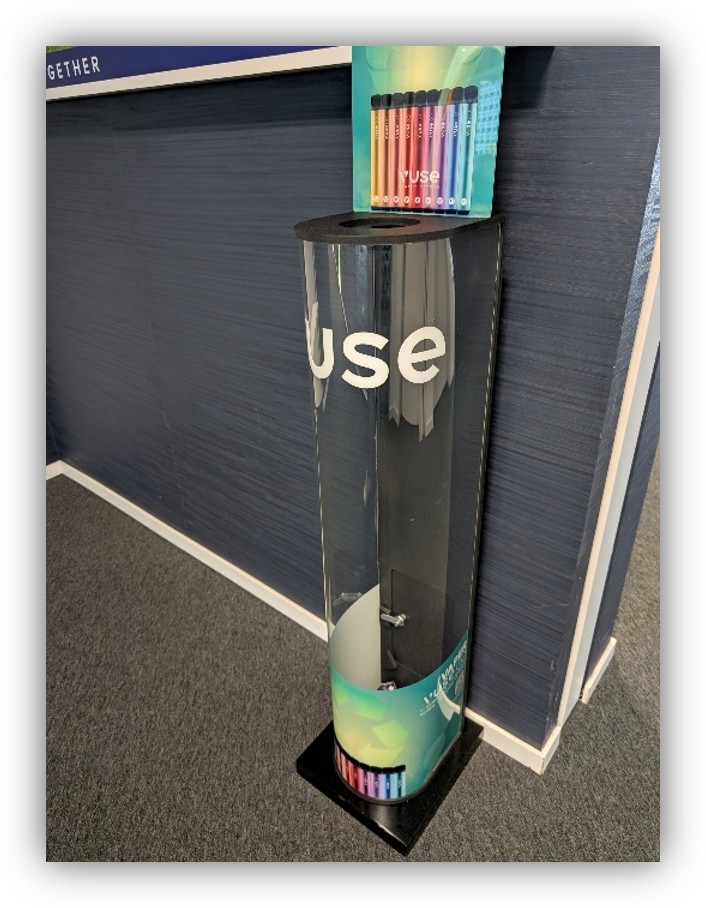 Dispensador transparente para la recogida y reutilización de dispositivos electrónicos, situado en una oficina y acompañado de varios dispositivos de colores en la parte superior e inferior.