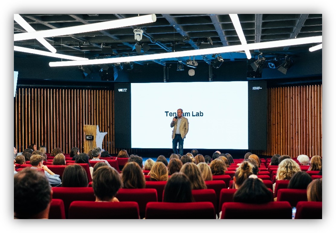 Ponente realizando una presentación ante una audiencia en un auditorio, con una pantalla de gran formato al fondo.