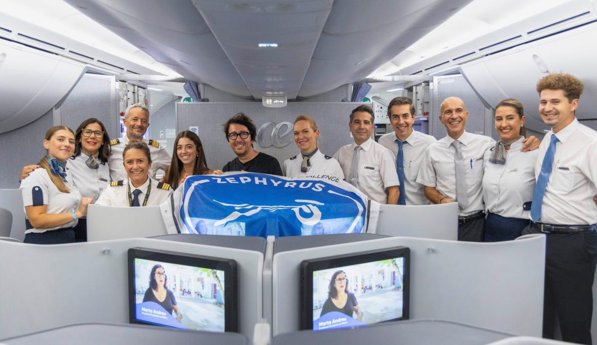 Grupo de personal uniformado posando dentro de la cabina de un avión, detrás de una bandera azul con el texto 'ZEPHYRUS'. En primer plano se ven asientos de clase ejecutiva con pantallas individuales mostrando contenido audiovisual. El interior del avión es moderno, con iluminación blanca y compartimentos superiores cerrados.