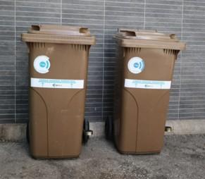 Dos contenedores marrones para residuos orgánicos, colocados uno al lado del otro sobre un suelo de cemento, frente a una pared gris con azulejos. Cada contenedor tiene una etiqueta blanca con un logotipo circular azul y texto que indica “Materia orgánica biodegradable”.