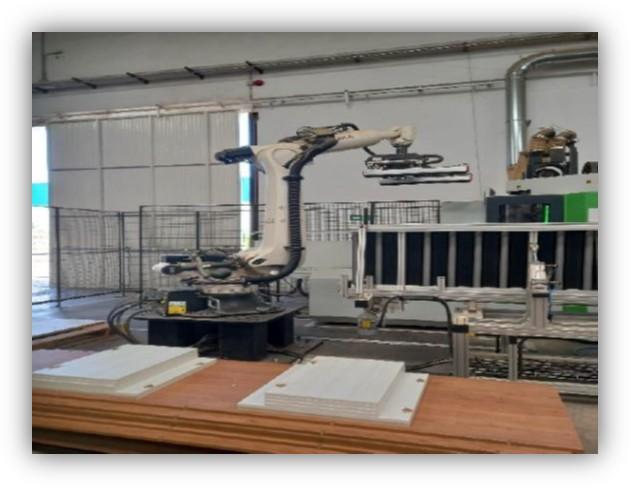 Brazo robótico industrial tomando o colocando piezas rectangulares blancas sobre una mesa de trabajo dentro de una nave de producción, con maquinaria y vallas de seguridad alrededor.