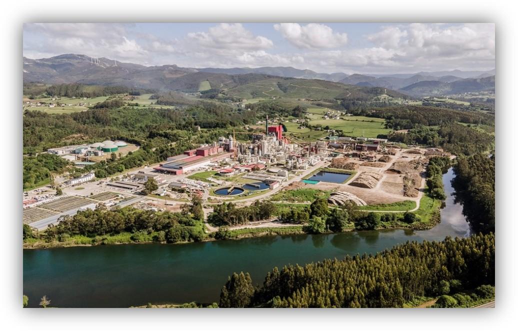 Vista aérea de una gran planta industrial rodeada de campos verdes, bosques, montañas y un río.