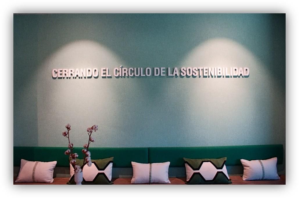 Pared verde con la frase ‘Cerrando el círculo de la sostenibilidad’ y un banco con varios cojines decorativos.