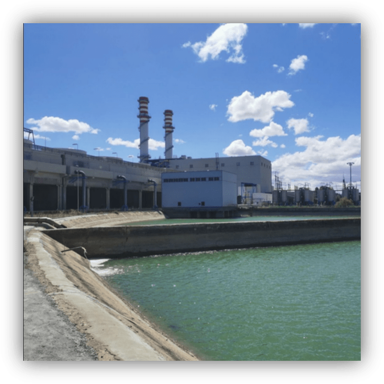 Vista de una planta industrial junto a un canal de agua verde, con dos chimeneas altas al fondo bajo un cielo azul con nubes.
