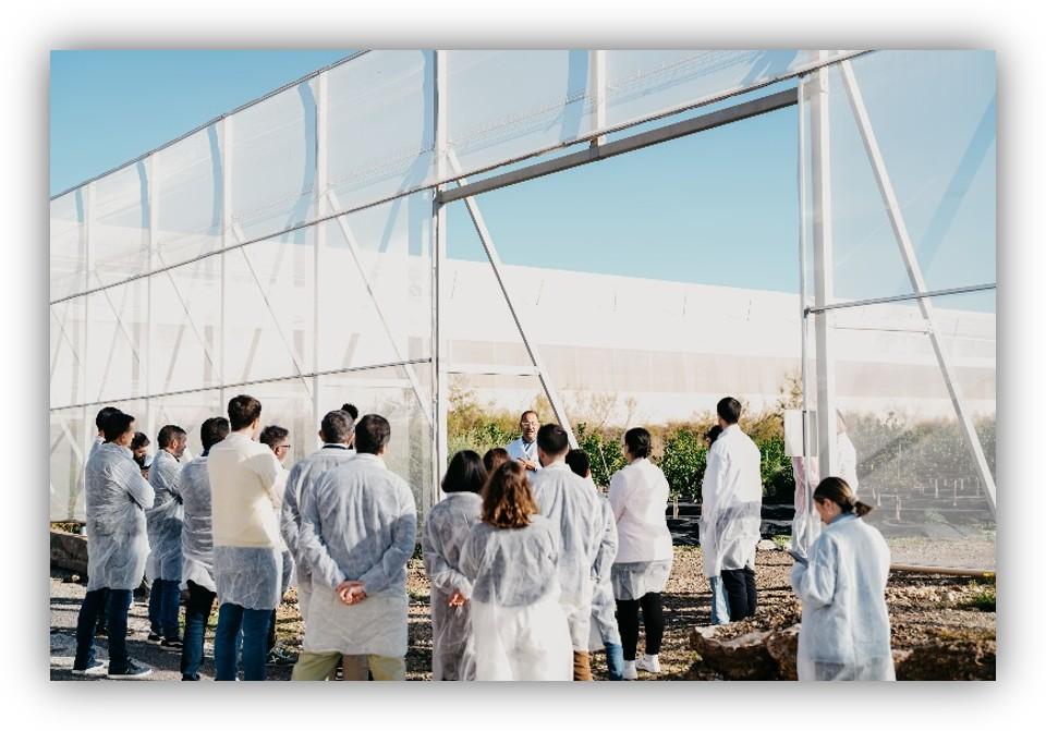 Grupo de personas con batas desechables escuchando una explicación en el exterior, frente a una estructura metálica y transparente.