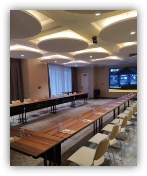 Sala de reuniones preparada para una conferencia, con mesas en forma de U, sillas alineadas y una pantalla grande encendida al fondo.