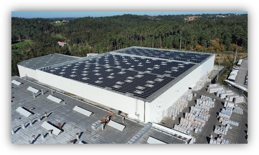 Vista aérea de una nave industrial con paneles solares en la cubierta, rodeada de arbolado y zonas de almacenamiento exterior.
