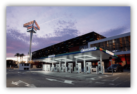 Estación de recarga de vehículos eléctricos frente a un complejo comercial al atardecer.