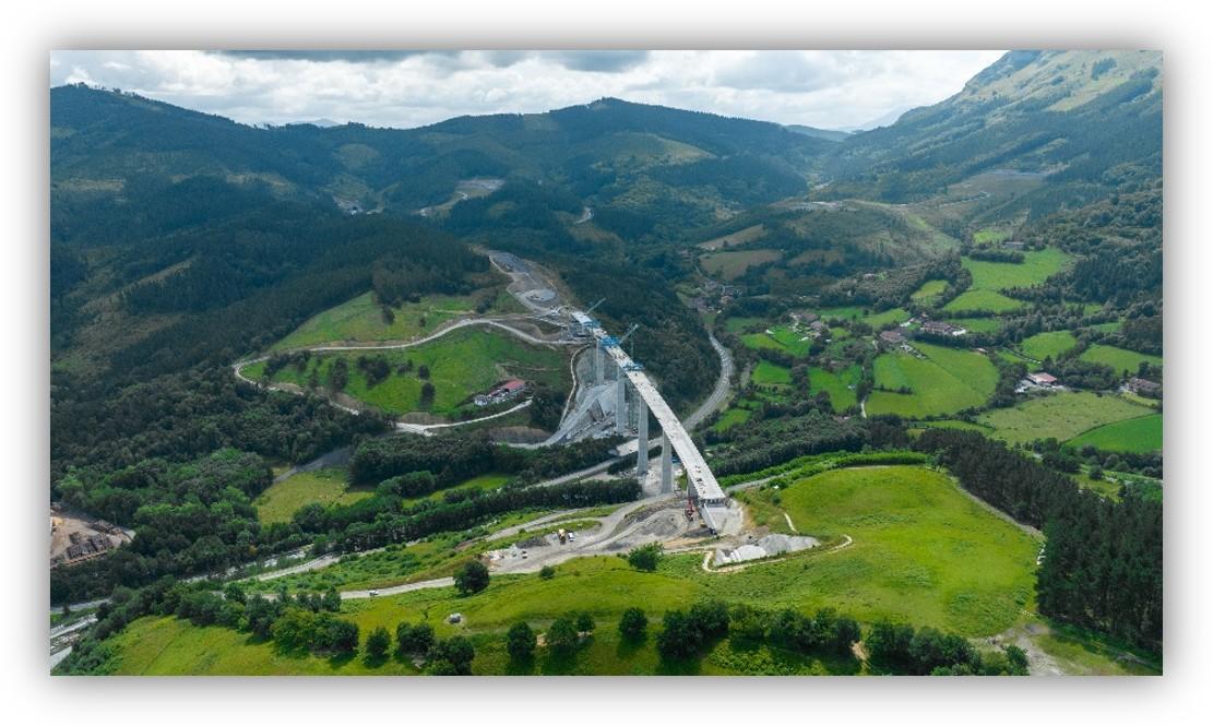 Vista aérea de un viaducto en construcción que atraviesa un valle verde, rodeado de montañas, bosques y praderas.