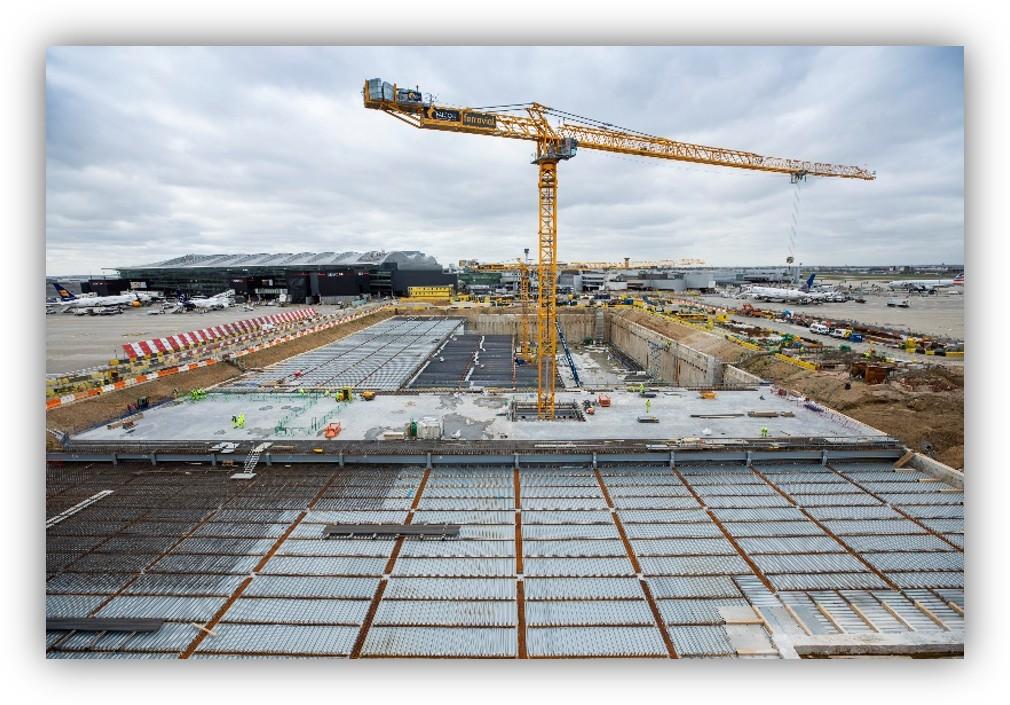 Obras de construcción en un aeropuerto con una gran grúa torre, una excavación de gran tamaño y varias aeronaves visibles al fondo.