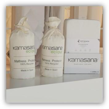 Tres botes blancos y dos cajas de protectores de colchón Kamasana sobre una mesa blanca.