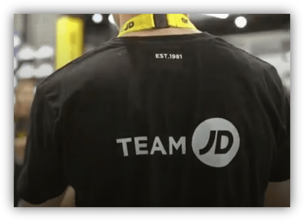 Parte trasera de una camiseta negra con el logotipo "Team JD" en un entorno comercial o logístico.