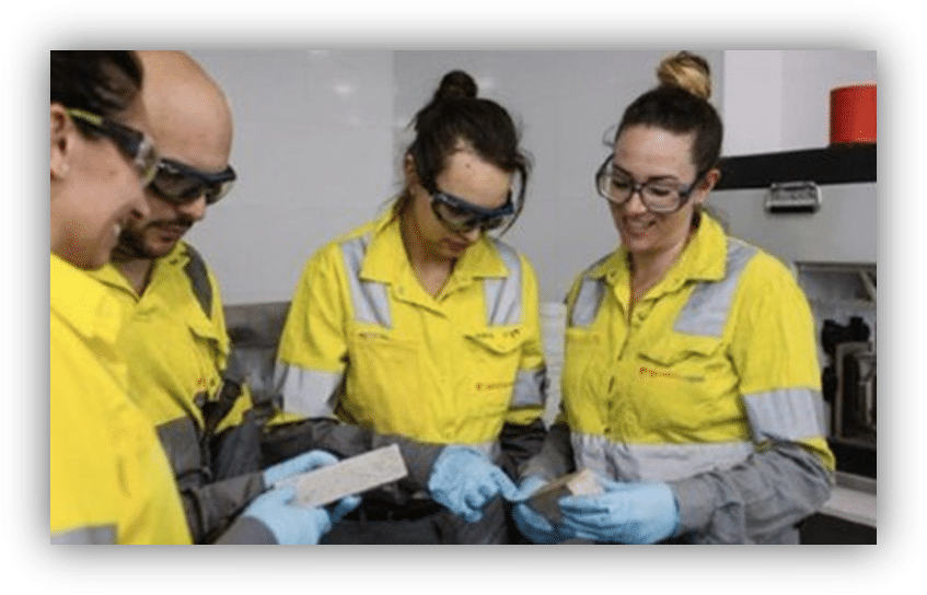 Cuatro personas con ropa de protección de alta visibilidad, gafas de seguridad y guantes examinan varias muestras en un laboratorio.