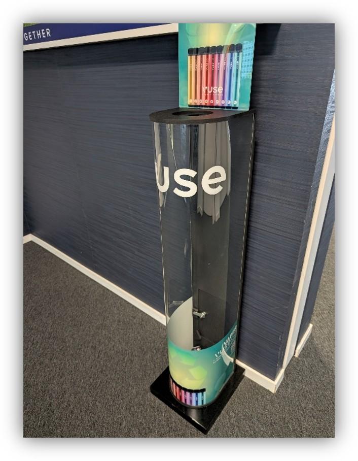 Dispensador transparente para la recogida y reutilización de dispositivos electrónicos, situado en una oficina y acompañado de varios dispositivos de colores en la parte superior e inferior.