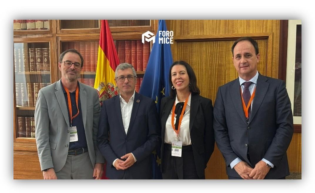 Cuatro personas posan en una biblioteca frente a las banderas de España y de la Unión Europea durante un encuentro institucional.
