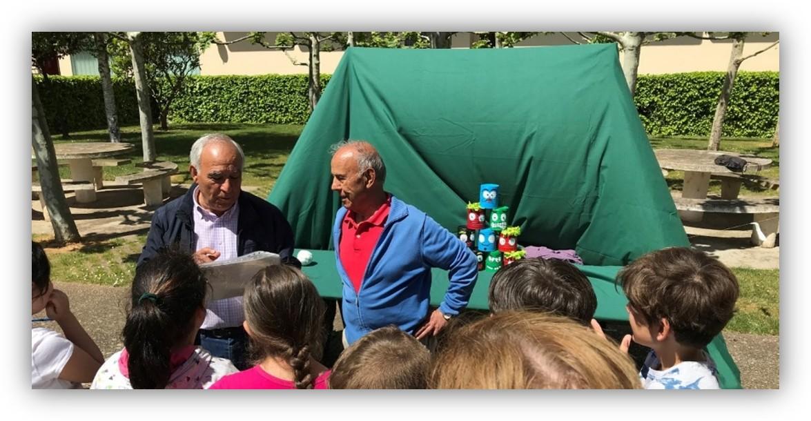 Grupo de escolares escuchando una explicación al aire libre durante una actividad educativa sobre reciclaje y medio ambiente, con materiales reutilizados expuestos sobre una mesa.
