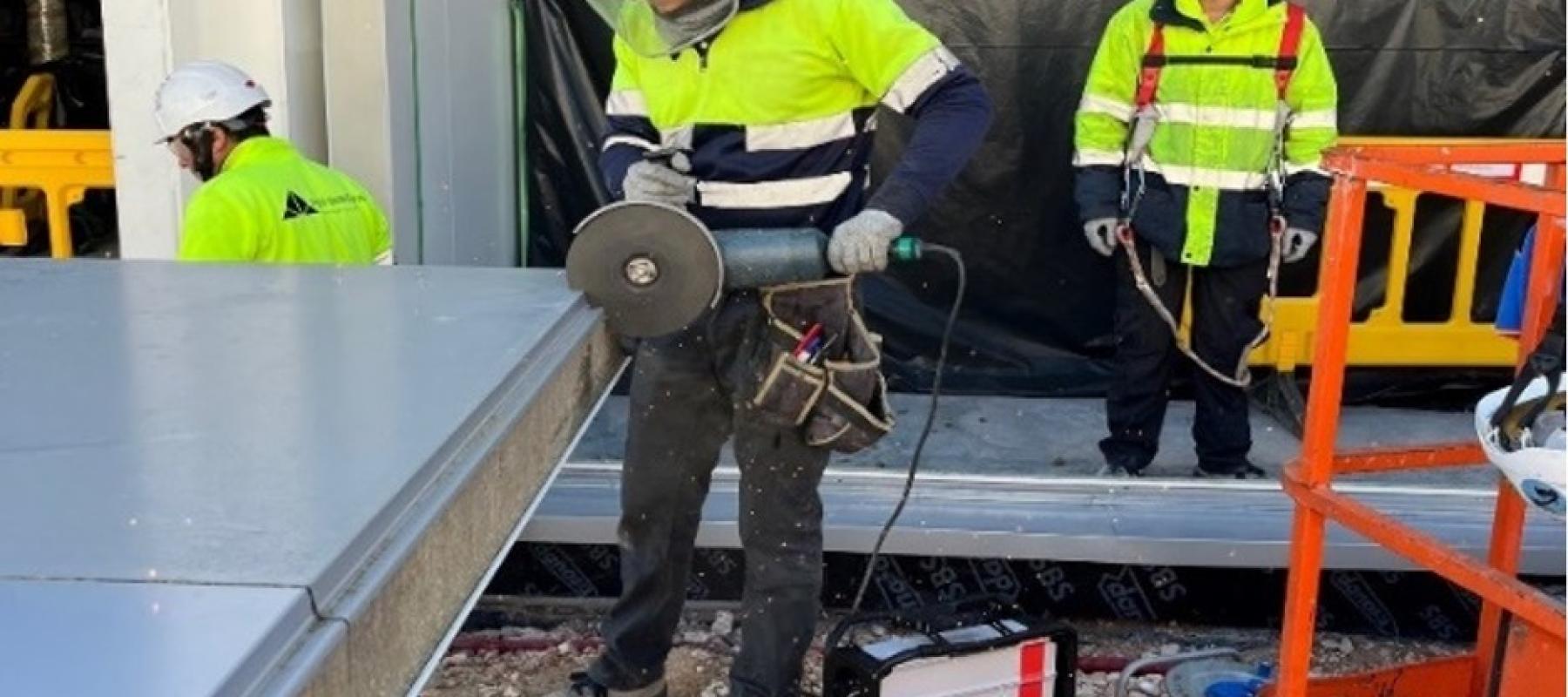 Un trabajador de construcción está utilizando una herramienta eléctrica tipo radial para cortar una pieza metálica en el borde de una estructura. Lleva equipo de protección personal, incluyendo casco, gafas, guantes y chaleco reflectante. En el suelo hay una máquina conectada por cable a la herramienta. Al fondo se observan otros trabajadores con ropa de alta visibilidad, una barrera amarilla y una estructura cubierta con plástico negro. También hay una plataforma elevadora naranja parcialmente visible en e