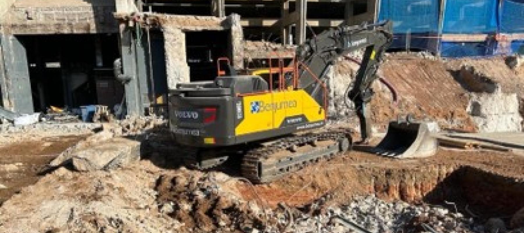 Excavadora de orugas Volvo, color amarillo y negro, con el logotipo de la empresa Benjumea en el costado, trabajando en una obra de demolición y excavación. El terreno está lleno de escombros y tierra removida, y al fondo se observan estructuras parcialmente demolidas y edificios en construcción con andamios y lonas azules.