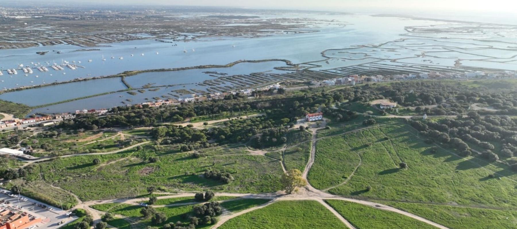 Vista aérea de una zona costera con amplias áreas verdes en primer plano, caminos de tierra que se cruzan y una pequeña edificación blanca y roja entre los árboles. Al fondo se observan marismas, canales de agua y un puerto con embarcaciones, todo bajo un cielo despejado.
