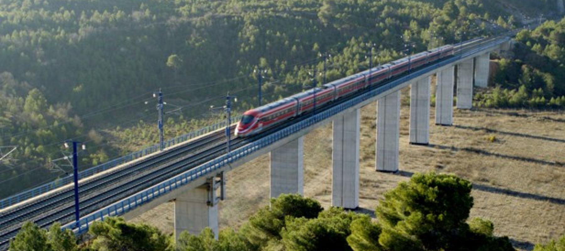 La imagen muestra un tren de alta velocidad circulando sobre un viaducto elevado en una zona montañosa. El tren es de color rojo con detalles en gris y está compuesto por varios vagones aerodinámicos. El viaducto está sostenido por pilares de hormigón altos y atraviesa un paisaje natural con abundante vegetación, principalmente pinos, en el fondo y en las laderas. El cielo no es visible, pero la iluminación indica que es de día, con luz natural clara.