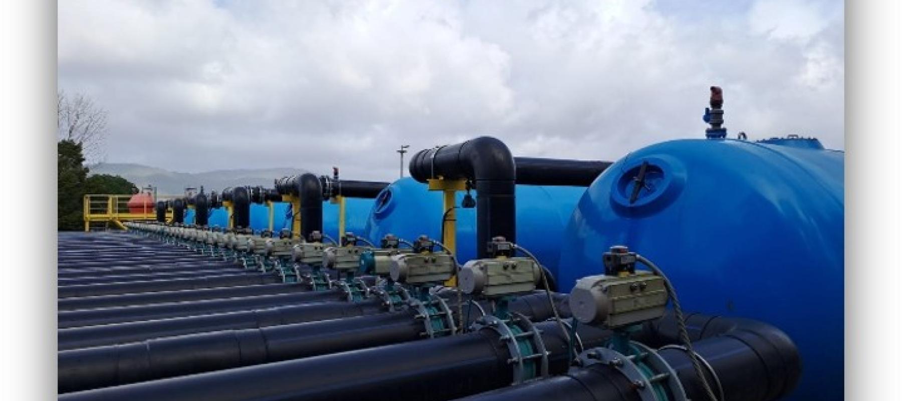 Instalación industrial con grandes tuberías negras y depósitos azules alineados al aire libre bajo un cielo nublado.