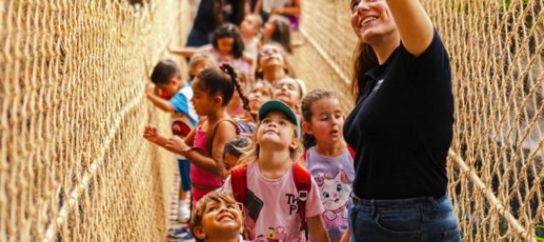 Un adulto y varios niños cruzan un puente colgante de cuerdas en un entorno natural. El adulto señala hacia arriba y los niños observan con curiosidad, rodeados de vegetación y rocas.