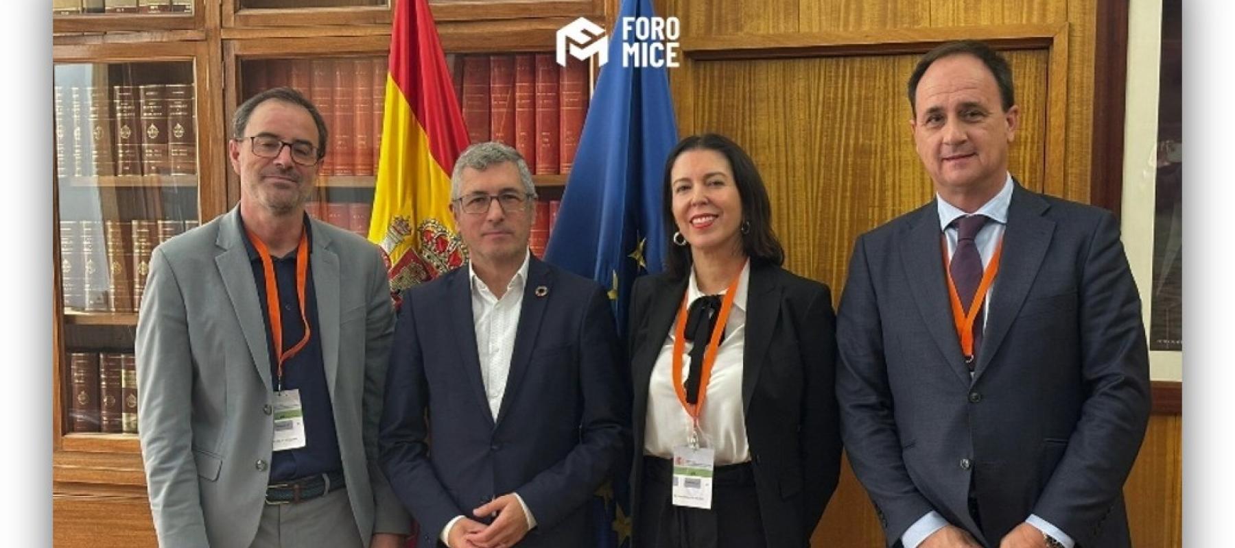 Cuatro personas posan en una biblioteca frente a las banderas de España y de la Unión Europea durante un encuentro institucional.