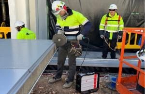 Un trabajador de construcción está utilizando una herramienta eléctrica tipo radial para cortar una pieza metálica en el borde de una estructura. Lleva equipo de protección personal, incluyendo casco, gafas, guantes y chaleco reflectante. En el suelo hay una máquina conectada por cable a la herramienta. Al fondo se observan otros trabajadores con ropa de alta visibilidad, una barrera amarilla y una estructura cubierta con plástico negro. También hay una plataforma elevadora naranja parcialmente visible en e