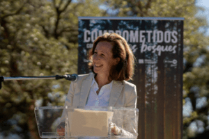 Persona hablando en un atril con micrófono durante un evento al aire libre, con un cartel de fondo que dice 'Comprometidos con los bosques