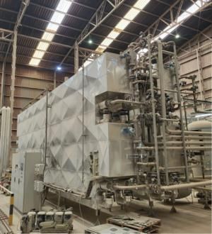 Equipo industrial de gran tamaño instalado en el interior de una nave, con estructura metálica recubierta por paneles de acero inoxidable con diseño geométrico. El sistema incluye múltiples tuberías, válvulas y componentes de control, así como un cuadro eléctrico en la parte frontal. El techo de la nave presenta vigas metálicas y paneles translúcidos que permiten la entrada de luz.