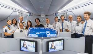 Grupo de personal uniformado posando dentro de la cabina de un avión, detrás de una bandera azul con el texto 'ZEPHYRUS'. En primer plano se ven asientos de clase ejecutiva con pantallas individuales mostrando contenido audiovisual. El interior del avión es moderno, con iluminación blanca y compartimentos superiores cerrados.