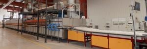 Vista interior de una planta industrial con un horno cerámico continuo de gran tamaño. El equipo está compuesto por una estructura metálica con paneles grises y naranjas, conductos superiores para extracción de gases y ventilación, y una cinta transportadora en la parte frontal para el movimiento de piezas. A la derecha se observan cuadros eléctricos y paneles de control con pantallas y botones. El suelo es de hormigón pulido y el entorno está bien iluminado, con paredes blancas y techo con vigas rojas